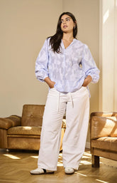 Woman wearing a light blue patterned blouse with a V neckline and long sleeves and white pants in a living room.