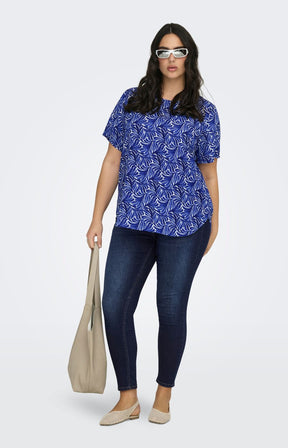 Woman wearing a blue patterned vica blouse and dark jeans, holding a beige tote bag on a white background