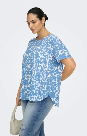 Woman wearing a blue and white floral patterned blouse and light wash jeans, holding a beige handbag on a light gray background