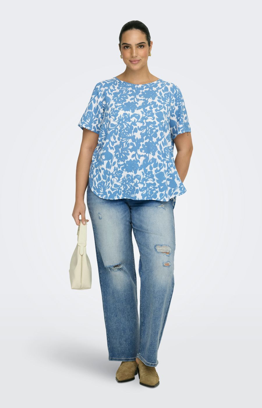 Woman wearing a blue and white floral patterned blouse and light wash jeans, holding a beige handbag on a light gray background