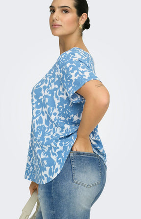 Woman wearing a blue and white floral patterned blouse and light wash jeans, holding a beige handbag on a light gray background 