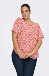 Woman wearing the Only Carmakoma Vica Blouse in Warm Tiles, a red and white patterned top on a light gray background