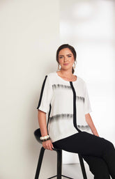 Woman wearing the Ora Blouse in White with Black, a white blouse with black patterns, with a round neckline, 1/2 length sleeves and a relaxed fit worn with black trousers against a plain background