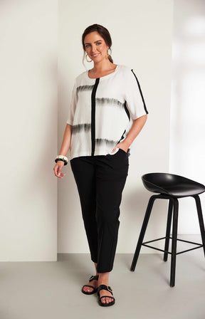 Woman wearing the Ora Blouse in White with Black, a white blouse with black patterns, with a round neckline, 1/2 length sleeves and a relaxed fit worn with black trousers against a plain background