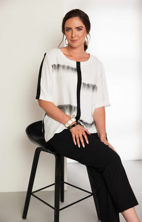 Woman wearing the Ora Blouse in White with Black, a white blouse with black patterns, with a round neckline, 1/2 length sleeves and a relaxed fit worn with black trousers against a plain background 