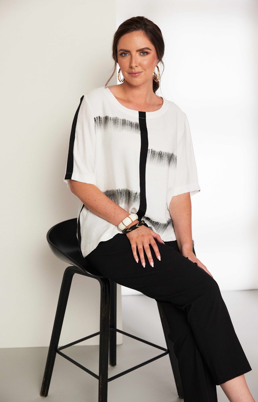 Woman wearing the Ora Blouse in White with Black, a white blouse with black patterns, with a round neckline, 1/2 length sleeves and a relaxed fit worn with black trousers against a plain background 