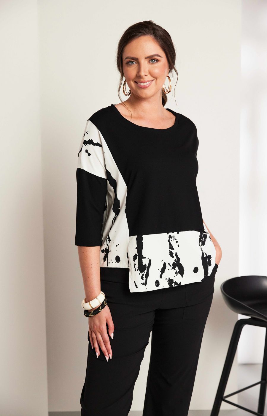 Woman wearing the Ora Contrasting Hem Top in Black, a black top with a contrasting side and hem with a round neckline, 1/2 length sleeves, cut out on the hem, pocket on the front with a plain background