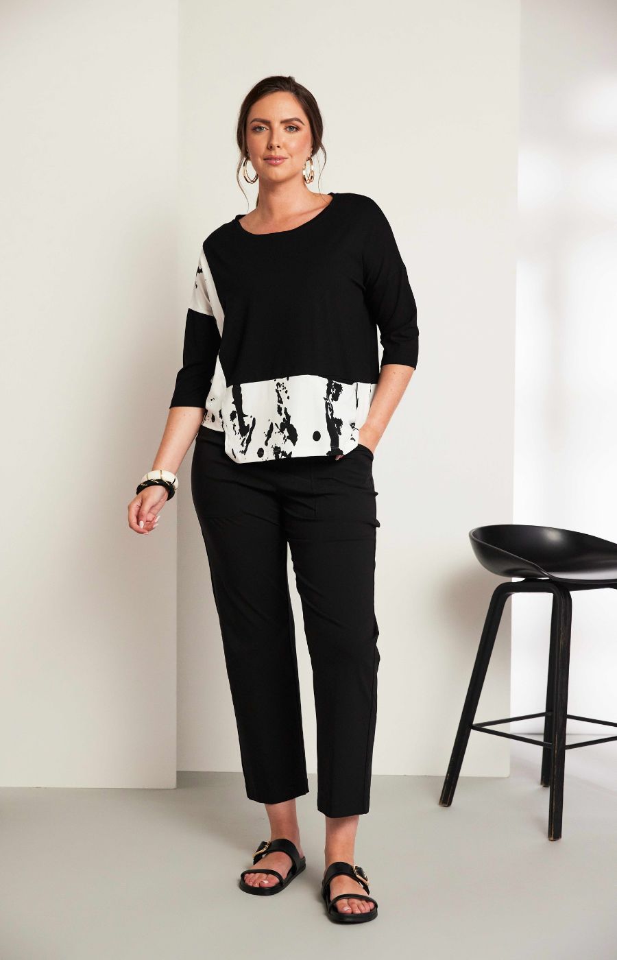 Woman wearing the Ora Contrasting Hem Top in Black, a black top with a contrasting side and hem with a round neckline, 1/2 length sleeves, cut out on the hem, pocket on the front with a plain background