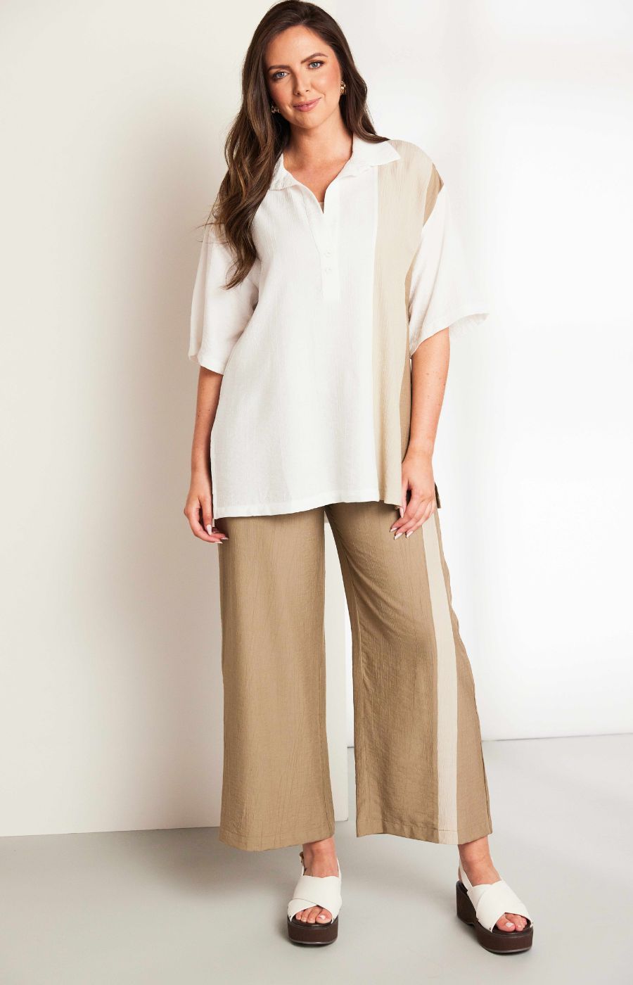 Woman wearing the Ora Crinkle Top with Collar in Beige, a white top with a beige side stripe down the front and back, a collard V neckline, short sleeves and side slits, worn with beige pants against a plain background