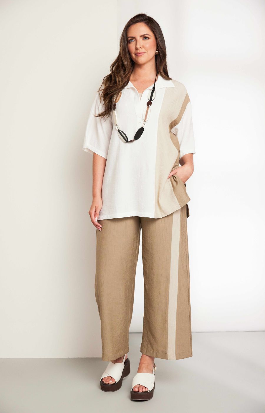 Woman wearing the Ora Crinkle Top with Collar in Beige, a white top with a beige side stripe down the front and back, a collard V neckline, short sleeves and side slits, worn with beige pants against a plain background 