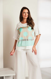 Woman wearing the Ora Dot T-Shirt with Turquoise, a lT-shirt with a round neckline and short sleeves  turquoise polka dots and sequin detailing and white trousers