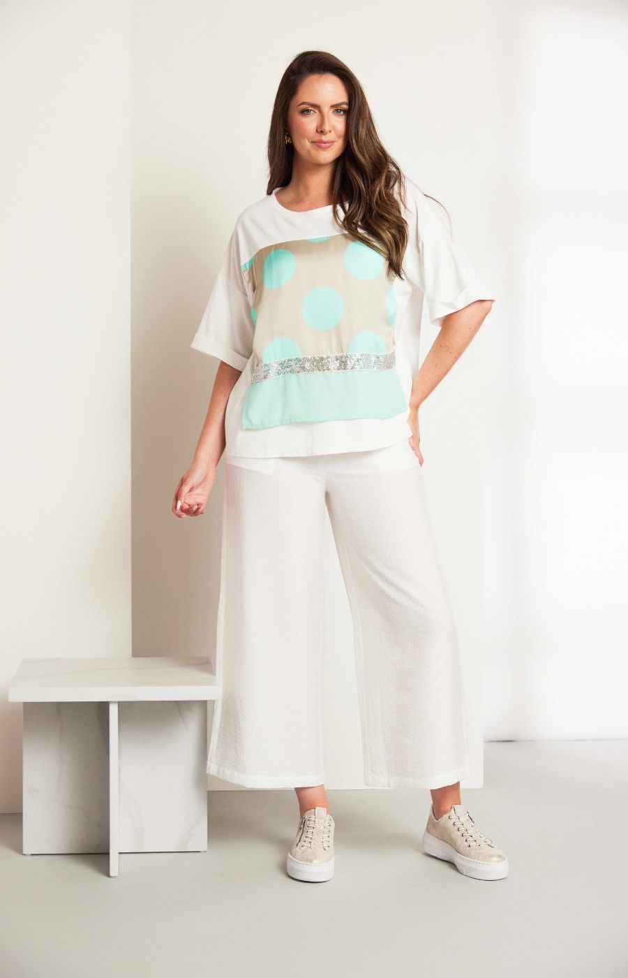 Woman wearing the Ora Dot T-Shirt with Turquoise, a lT-shirt with a round neckline and short sleeves  turquoise polka dots and sequin detailing and white trousers 