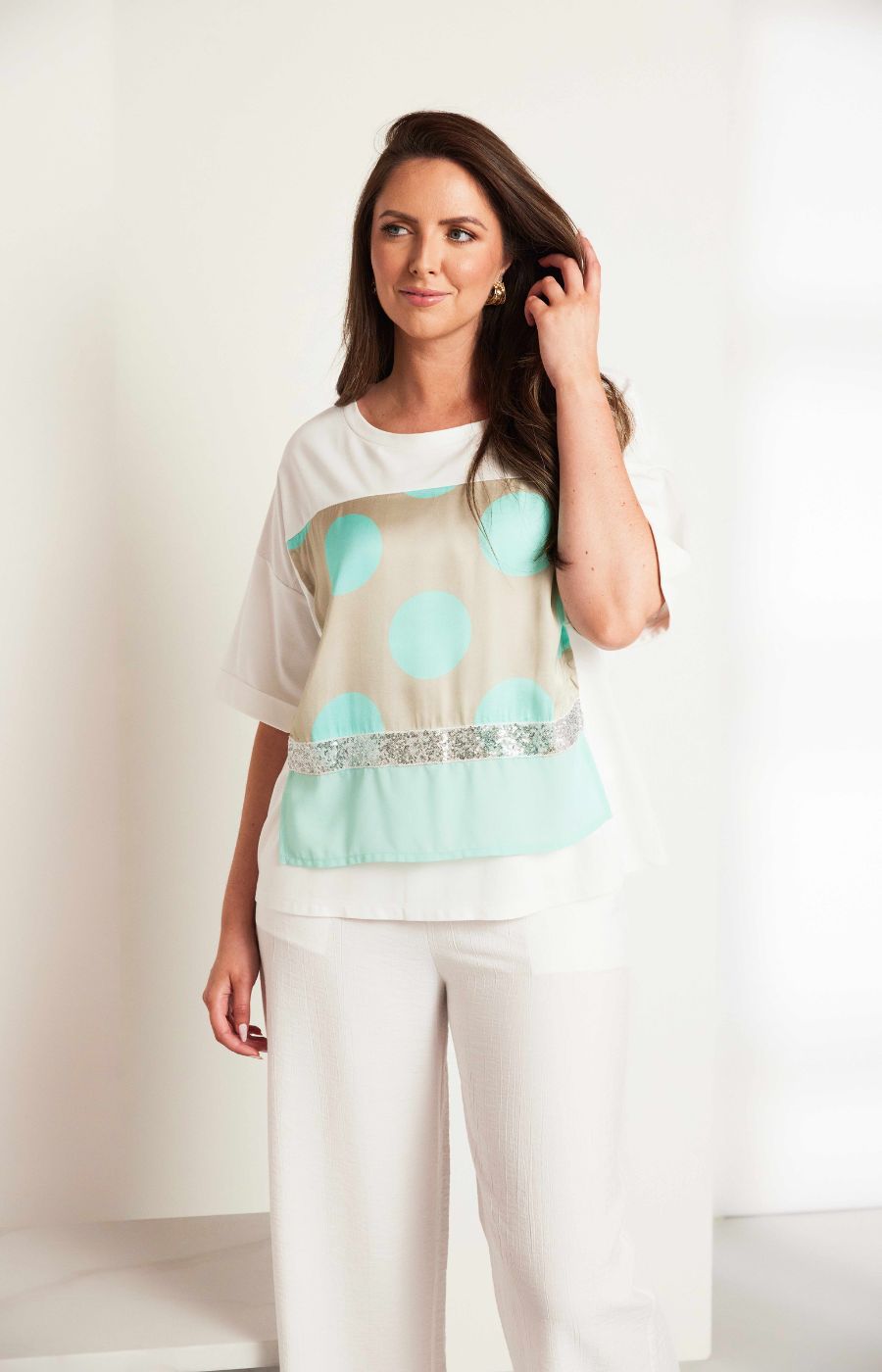 Woman wearing the Ora Dot T-Shirt with Turquoise, a lT-shirt with a round neckline and short sleeves  turquoise polka dots and sequin detailing and white trousers 