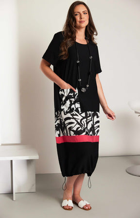 Woman wearing the Ora Dress in Black with Necklace, a black dress with a white, black and pink  design on the fron, front pocket, short sleeves and a round neckline against a white background