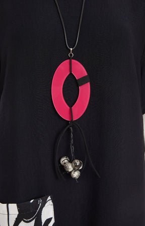 Woman wearing the Ora Dress in Black with Necklace, a black dress with a white, black and pink  design on the fron, front pocket, short sleeves and a round neckline against a white background 