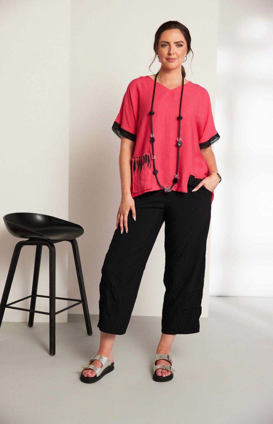 Woman wearing the Ora Feather Print Top in Pink, a pink blouse with black trim, front pocket,  V neckline and short sleeves and black trousers against a white background