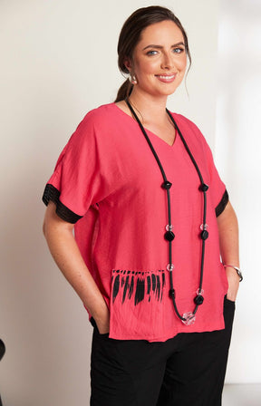 Woman wearing the Ora Feather Print Top in Pink, a pink blouse with black trim, front pocket,  V neckline and short sleeves and black trousers against a white background