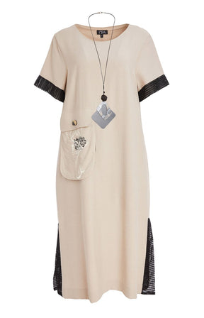 Woman wearing the Ora Front Pocket Dress in Beige, a beige midi dress with black mesh accents on the cuffs of the short sleeves and side slits, has a round neckline, large front pocket, discreet side pockets and necklace included in a minimal indoor setting  