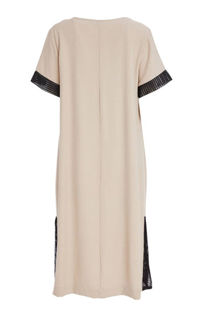 Woman wearing the Ora Front Pocket Dress in Beige, a beige midi dress with black mesh accents on the cuffs of the short sleeves and side slits, has a round neckline, large front pocket, discreet side pockets and necklace included in a minimal indoor setting 
