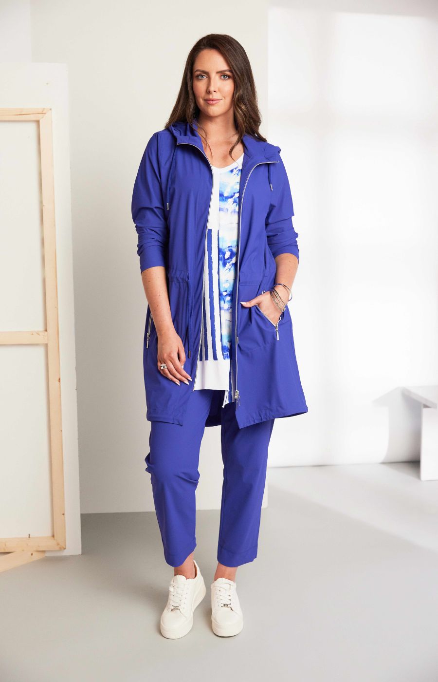 Woman wearing the Ora Hooded Jacket in Blue, with the blue Ora travel trousers a blue outfit with a white background  