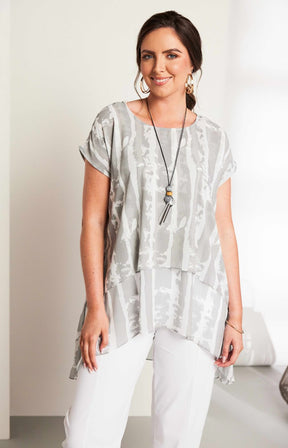 Woman wearing the Ora Layered Top in Grey, a slightly sheer, double layered top with a  light grey and white patterned top. Has a round neckline, short sleeves and a curved hem worn with white trousers against a plain background