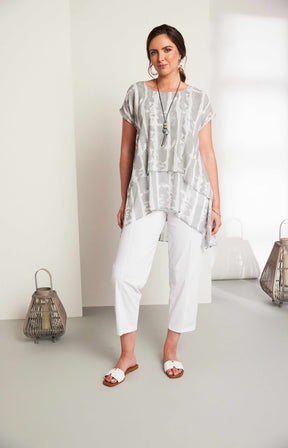 Woman wearing the Ora Layered Top in Grey, a slightly sheer, double layered top with a  light grey and white patterned top. Has a round neckline, short sleeves and a curved hem worn with white trousers against a plain background 