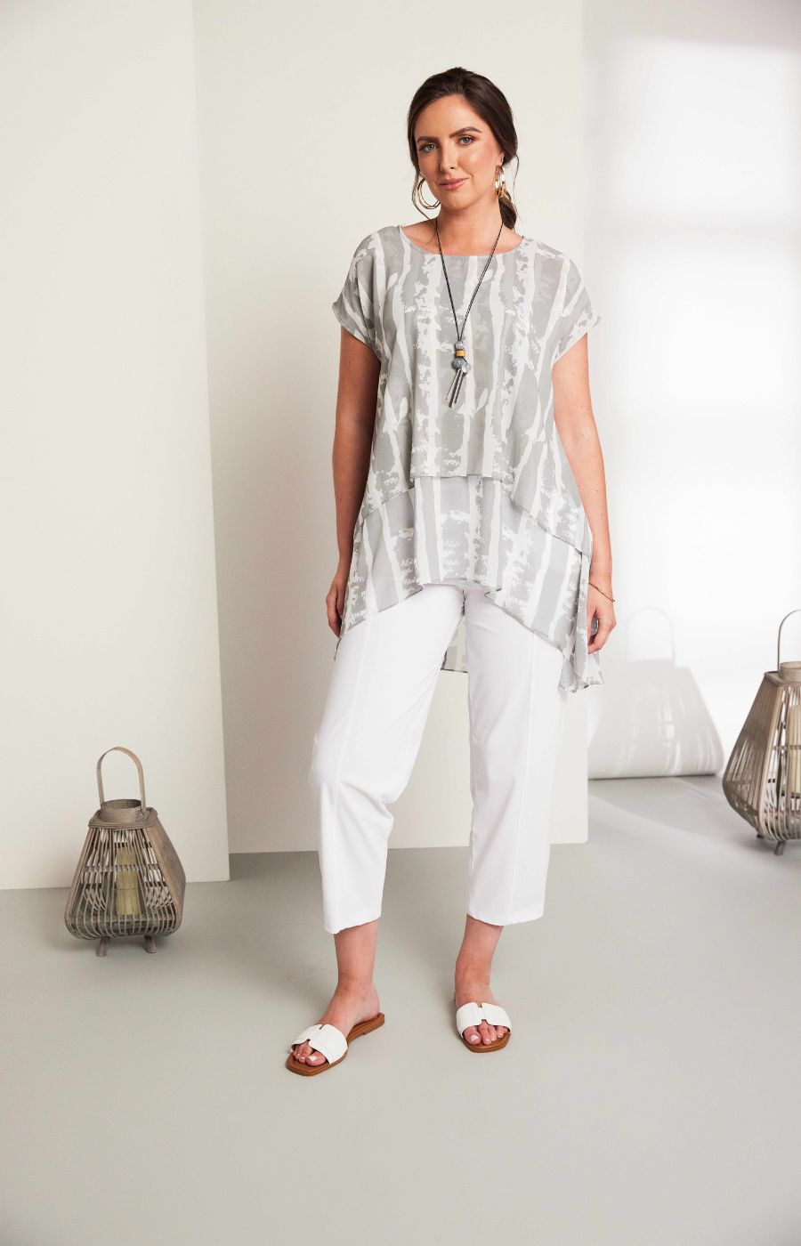 Woman wearing the Ora Layered Top in Grey, a slightly sheer, double layered top with a  light grey and white patterned top. Has a round neckline, short sleeves and a curved hem worn with white trousers against a plain background 