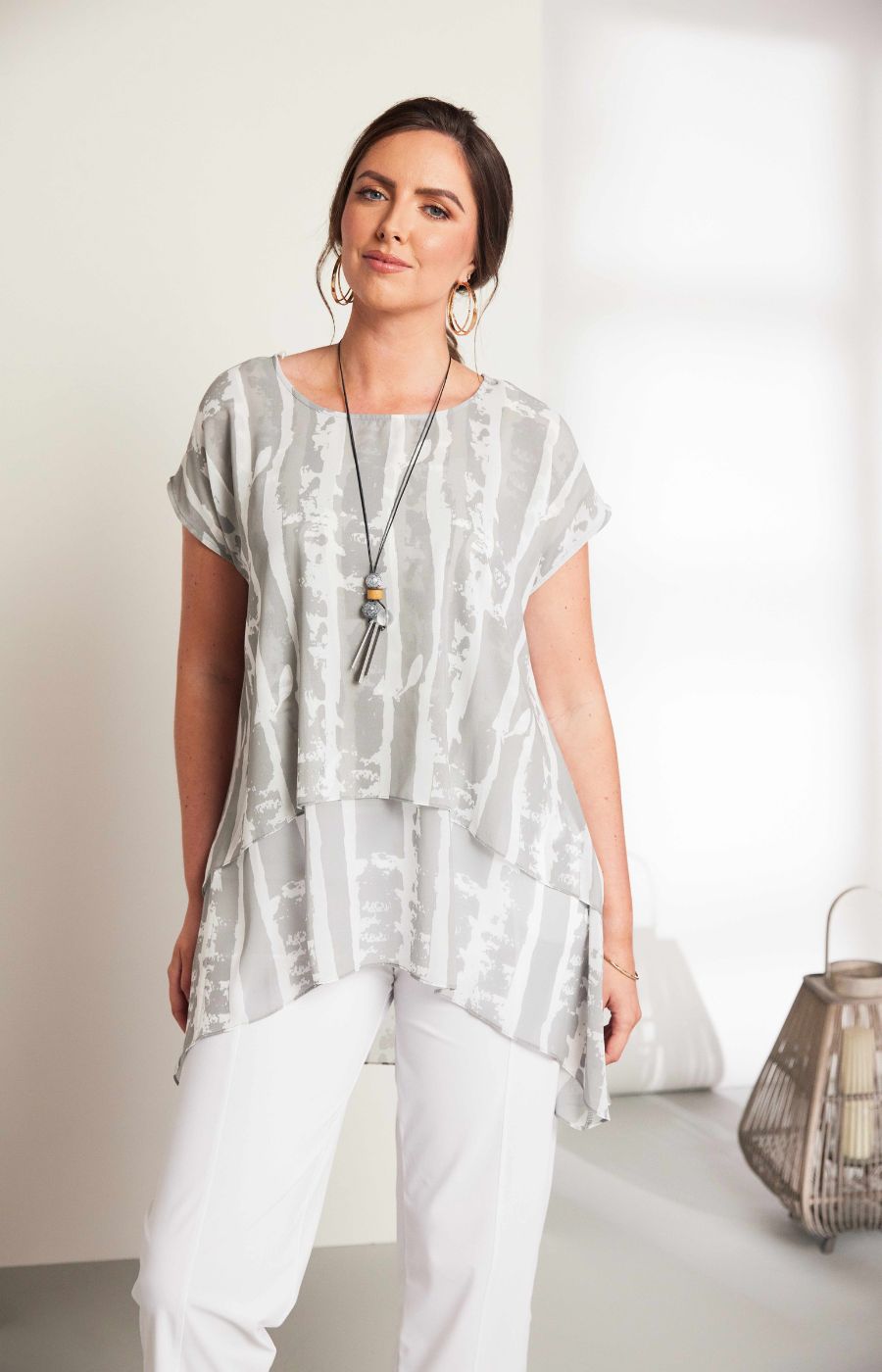 Woman wearing the Ora Layered Top in Grey, a slightly sheer, double layered top with a  light grey and white patterned top. Has a round neckline, short sleeves and a curved hem worn with white trousers against a plain background  