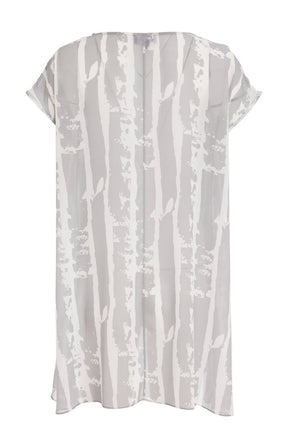 Woman wearing the Ora Layered Top in Grey, a slightly sheer, double layered top with a  light grey and white patterned top. Has a round neckline, short sleeves and a curved hem worn with white trousers against a plain background    