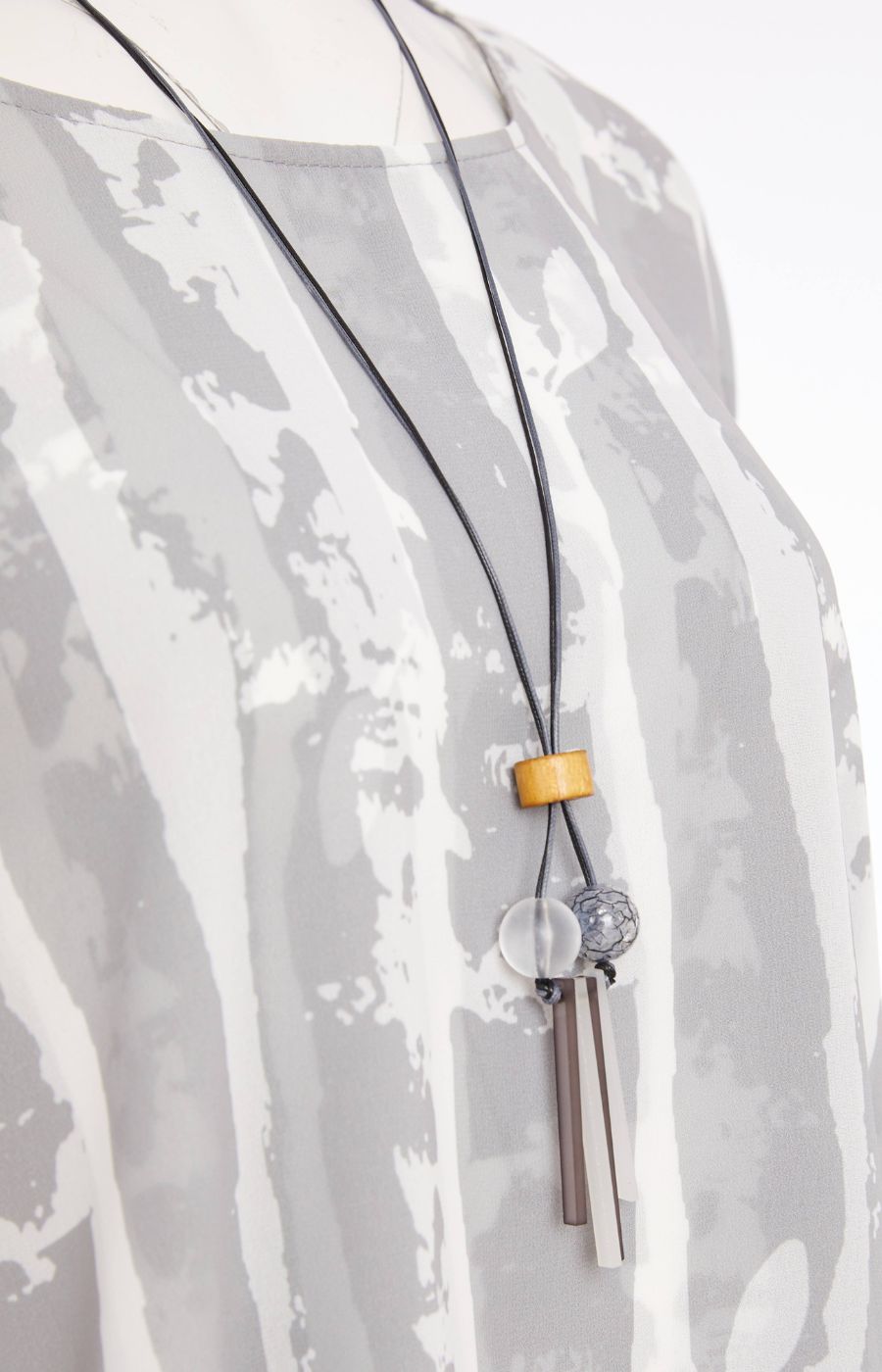 Woman wearing the Ora Layered Top in Grey, a slightly sheer, double layered top with a  light grey and white patterned top. Has a round neckline, short sleeves and a curved hem worn with white trousers against a plain background    