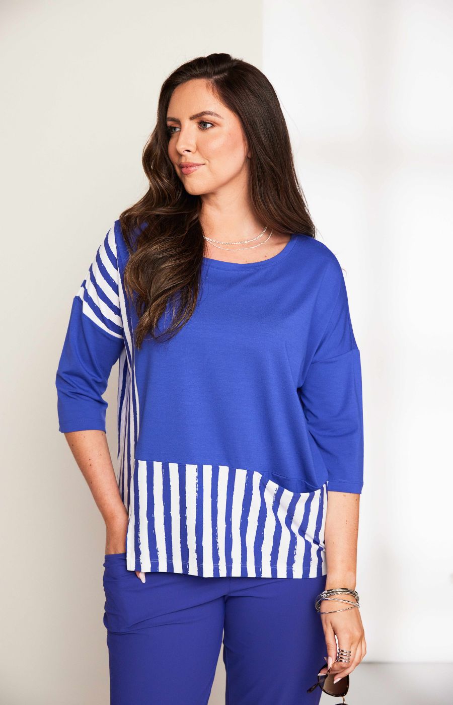 Woman wearing the Ora Striped Pocket Top in Blue, a blue top a round neckline and 3/4 length sleeves with white striped hem against a plain background