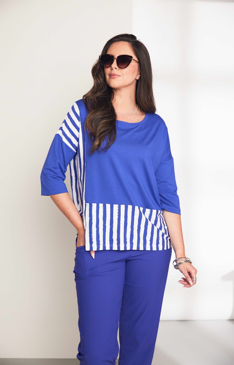 Woman wearing the Ora Striped Pocket Top in Blue, a blue top a round neckline and 3/4 length sleeves with white striped hem against a plain background  