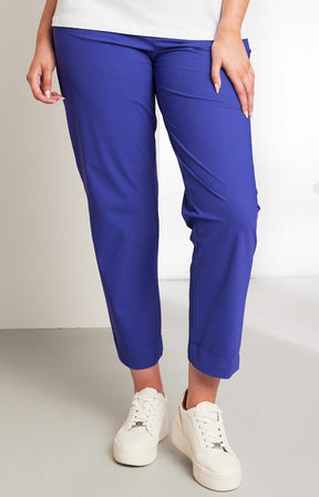 Women wearing the Ora Travel Trousers in Blue, a blue pull up style, full length trouser with an elasticated waistband and side pockets, worn with white sneakers on a plain background