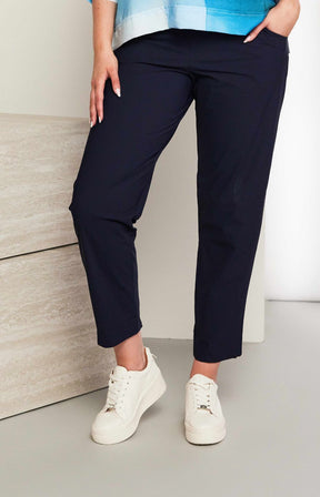 Women wearing the Ora Travel Trousers in Navy, a full length navy trouser with a straight leg, elasticated waistband and side pockets worn with white sneakers against a neutral background