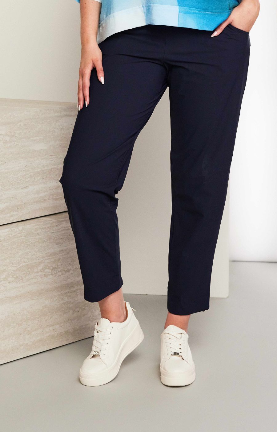 Women wearing the Ora Travel Trousers in Navy, a full length navy trouser with a straight leg, elasticated waistband and side pockets worn with white sneakers against a neutral background