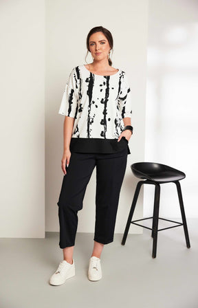 Woman wearing the Ora White and Black Stripe Top, a black and white messy stripe patterned top with 1/2 length sleeves, round neckline, relaxed fit and contrasting hem worn with black trousers. 