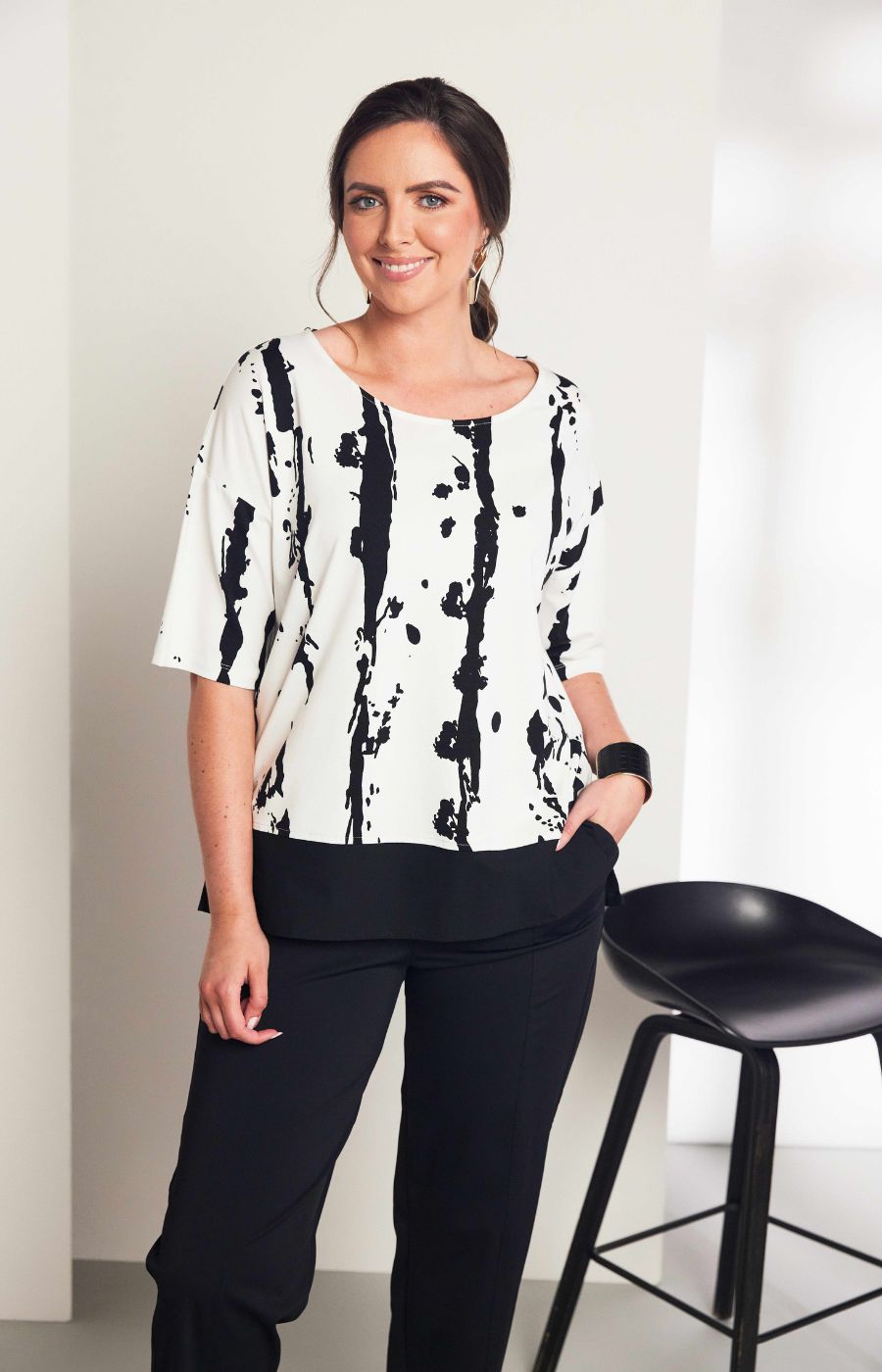 Woman wearing the Ora White and Black Stripe Top, a black and white messy stripe patterned top with 1/2 length sleeves, round neckline, relaxed fit and contrasting hem worn with black trousers. 