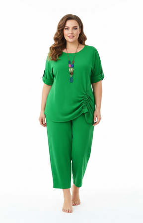 A woman standing against a plain white background wearing a green blouse with rolled-up sleeves and a gathered detail on one side, paired with matching green cropped pants. Accessorized with a long, colorful pendant necklace.