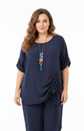 A woman standing against a plain white background wearing a navy blouse with rolled-up sleeves and a gathered detail on one side, paired with matching navy pants. Accessorized with a long, colorful pendant necklace.