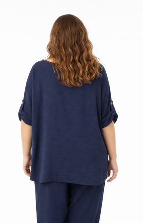 A woman standing against a plain white background wearing a navy blouse with rolled-up sleeves, paired with matching navy pants.