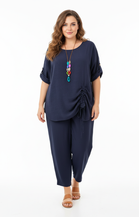 A woman standing against a plain white background wearing a navy blouse with rolled-up sleeves and a gathered detail on one side, paired with matching navy cropped pants. She is barefoot and accessorized with a long, colorful pendant necklace.