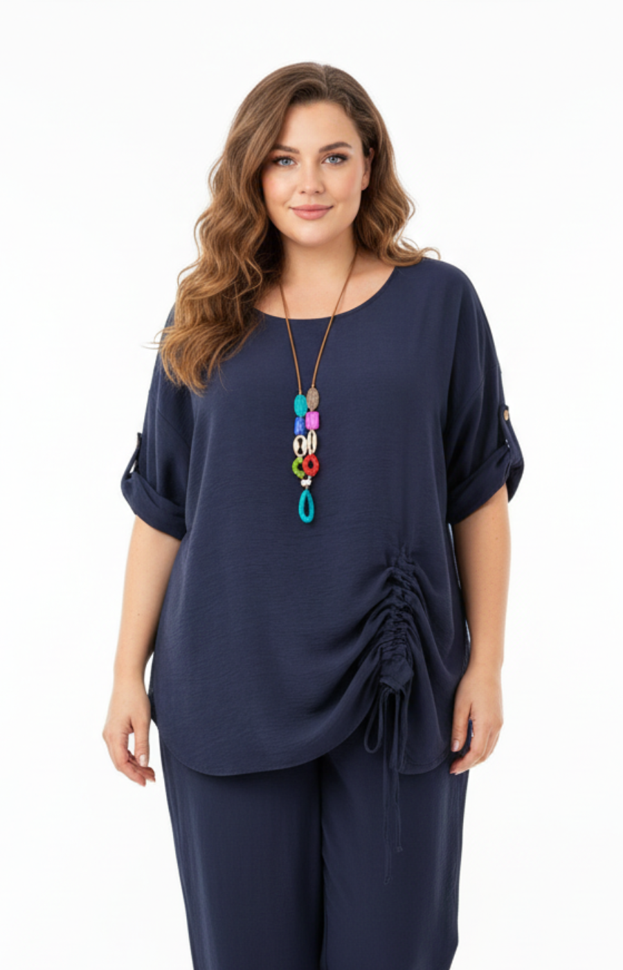 A woman standing against a plain white background wearing a navy blouse with rolled-up sleeves and a gathered detail on one side, paired with matching navy pants. Accessorized with a long, colorful pendant necklace.