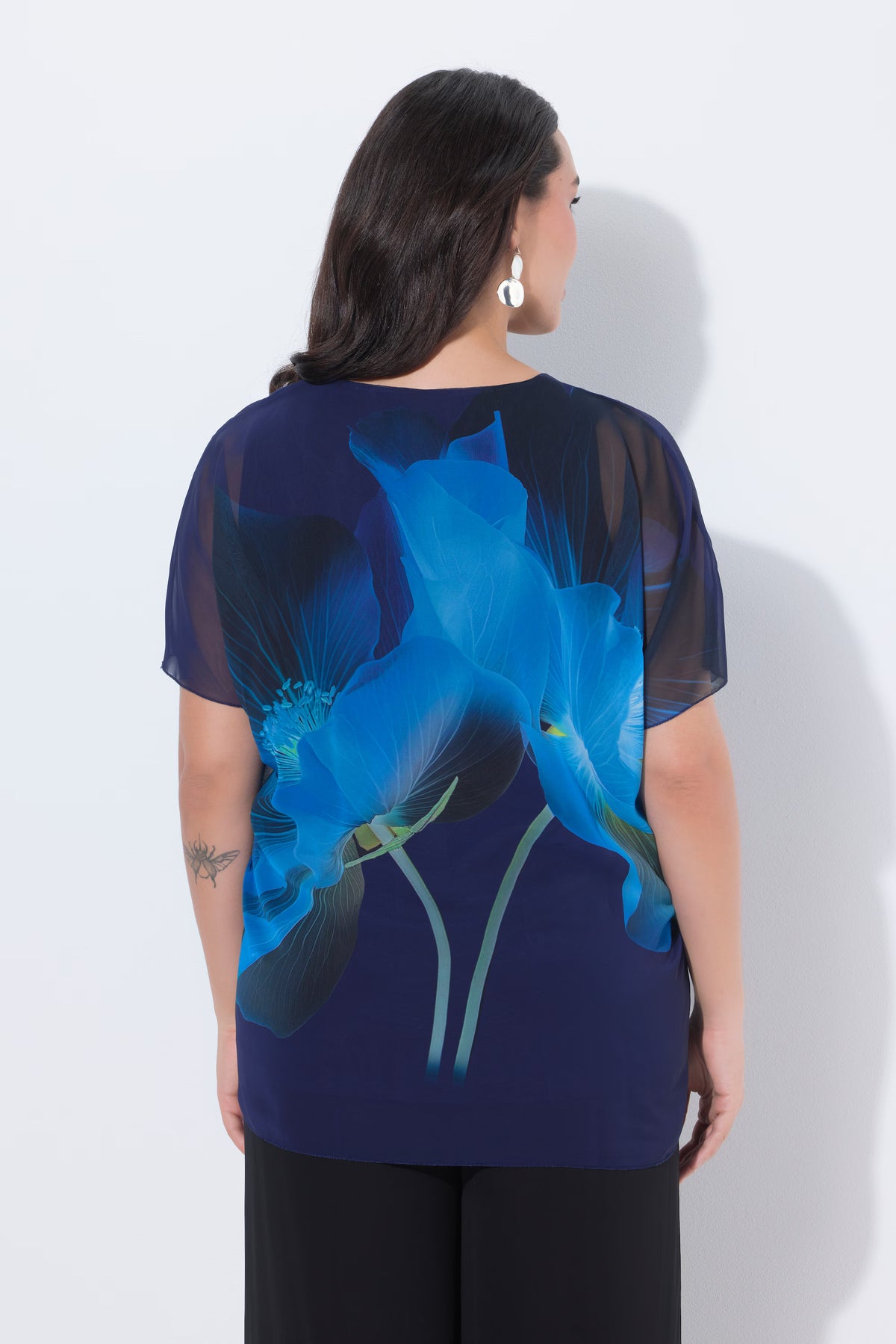 Woman wearing the Ulla Popken Floral Chiffon Blouse in Blue, a blue and navy floral blouse, with a floral design with rhinestone detailing, a round neckline and short sleeves against a white background