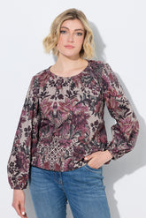 A woman wearing the Ulla Popken Floral Damask Blouse in Mauve, a long-sleeve floral blouse with a dusty rose, purple, and black pattern on a beige background, paired with blue jeans, against a light gray studio background.