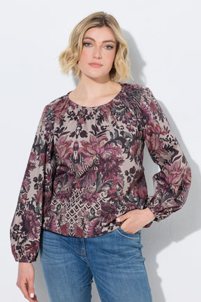 A woman wearing the Ulla Popken Floral Damask Blouse in Mauve, a long-sleeve floral blouse with a dusty rose, purple, and black pattern on a beige background, paired with blue jeans, against a light gray studio background.