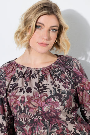 A woman with short blonde hair wearing the Ulla Popken Floral Damask Blouse in Mauve, a long-sleeve blouse with a dusty rose and purple floral print on a beige background, photographed against a light gray wall.
