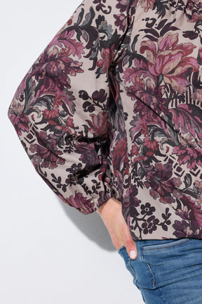 A close-up of a person wearing the Ulla Popken Floral Damask Blouse in Mauve,  a long-sleeve floral jacket with a beige base and dark purple, pink, and black botanical print, paired with blue jeans against a light gray background.