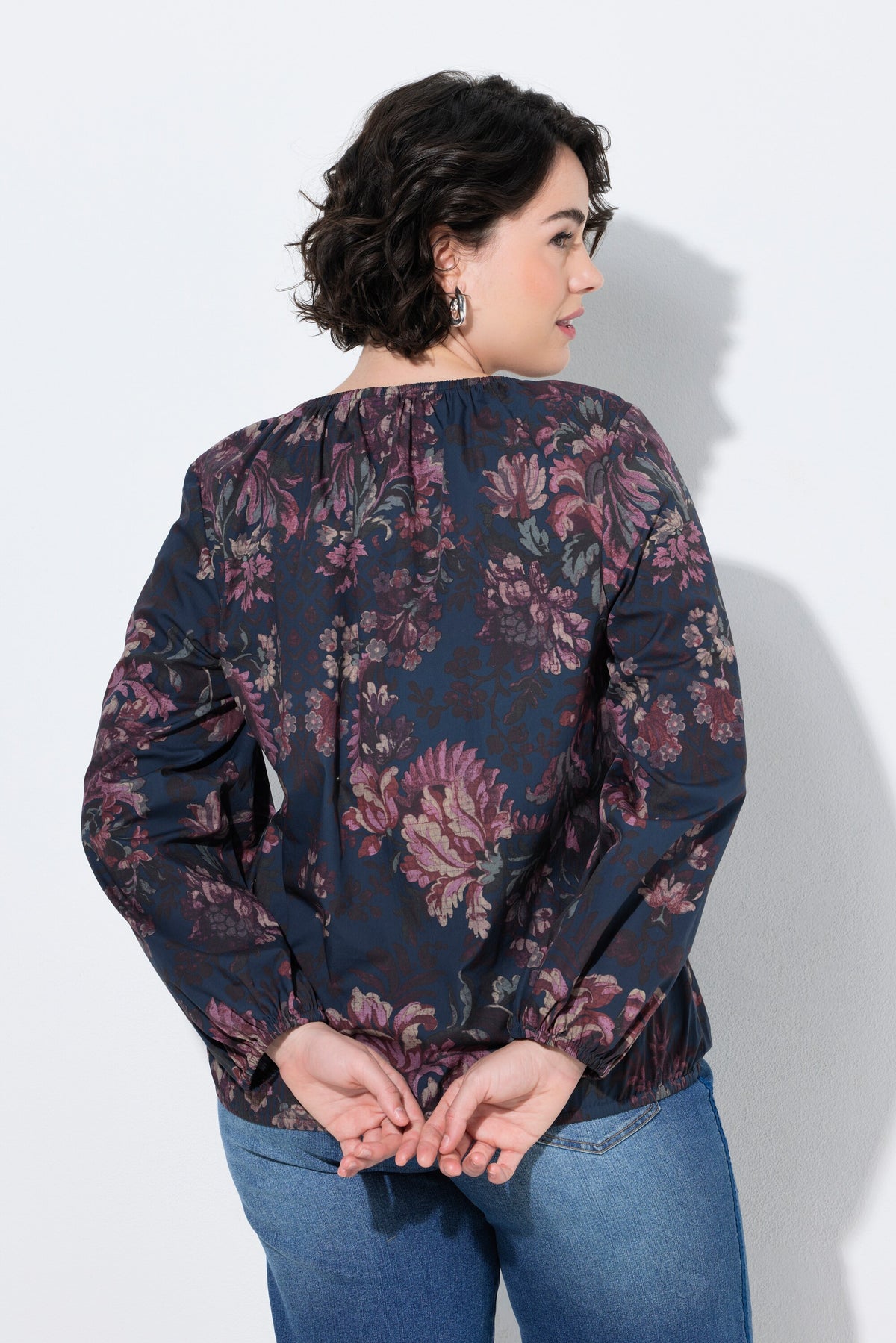A woman shown from the back wearing the Ulla Popken Floral Damask Blouse in Navy a dark navy floral blouse with long sleeves and blue jeans, standing against a light gray wall.