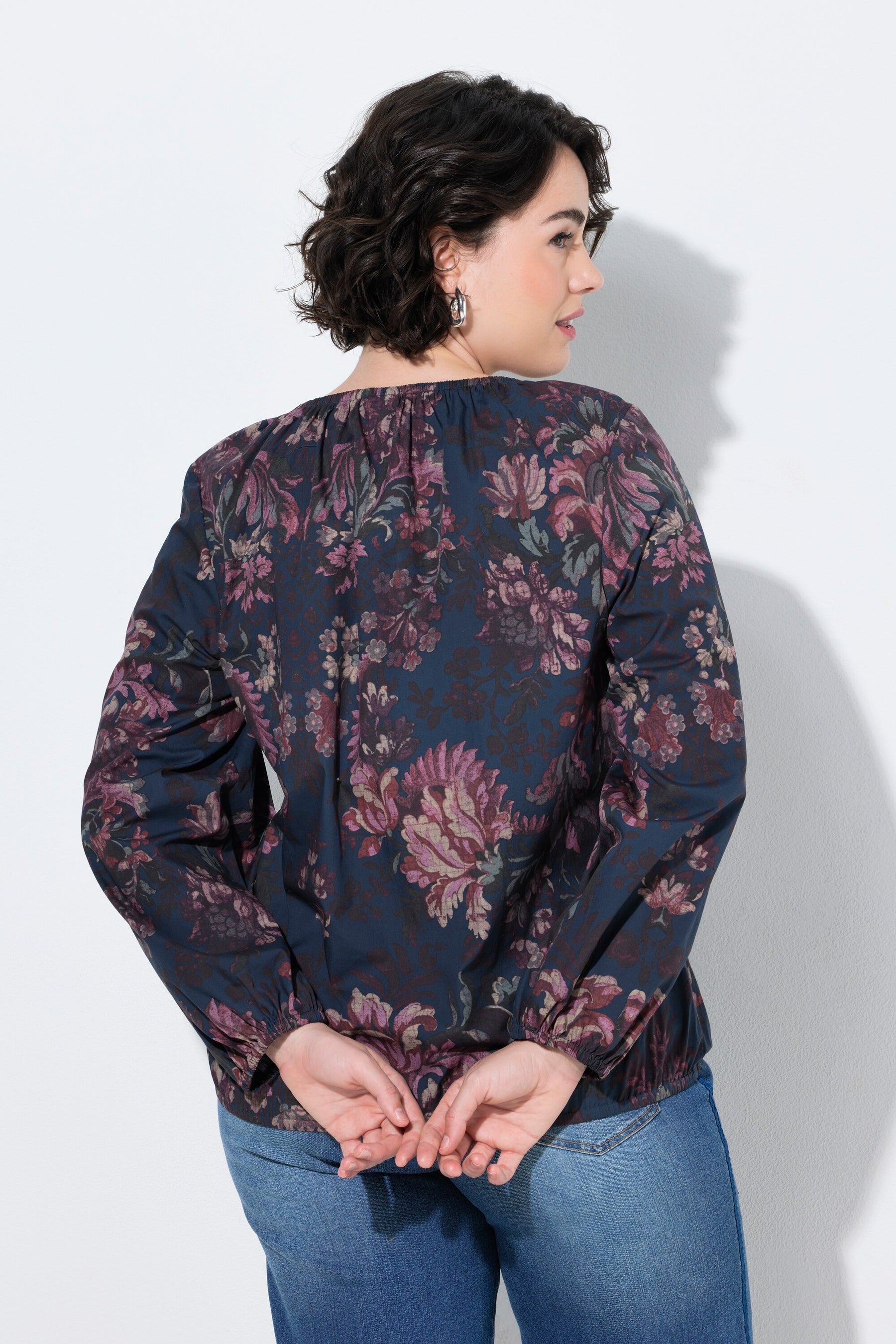 A woman shown from the back wearing the Ulla Popken Floral Damask Blouse in Navy a dark navy floral blouse with long sleeves and blue jeans, standing against a light gray wall.