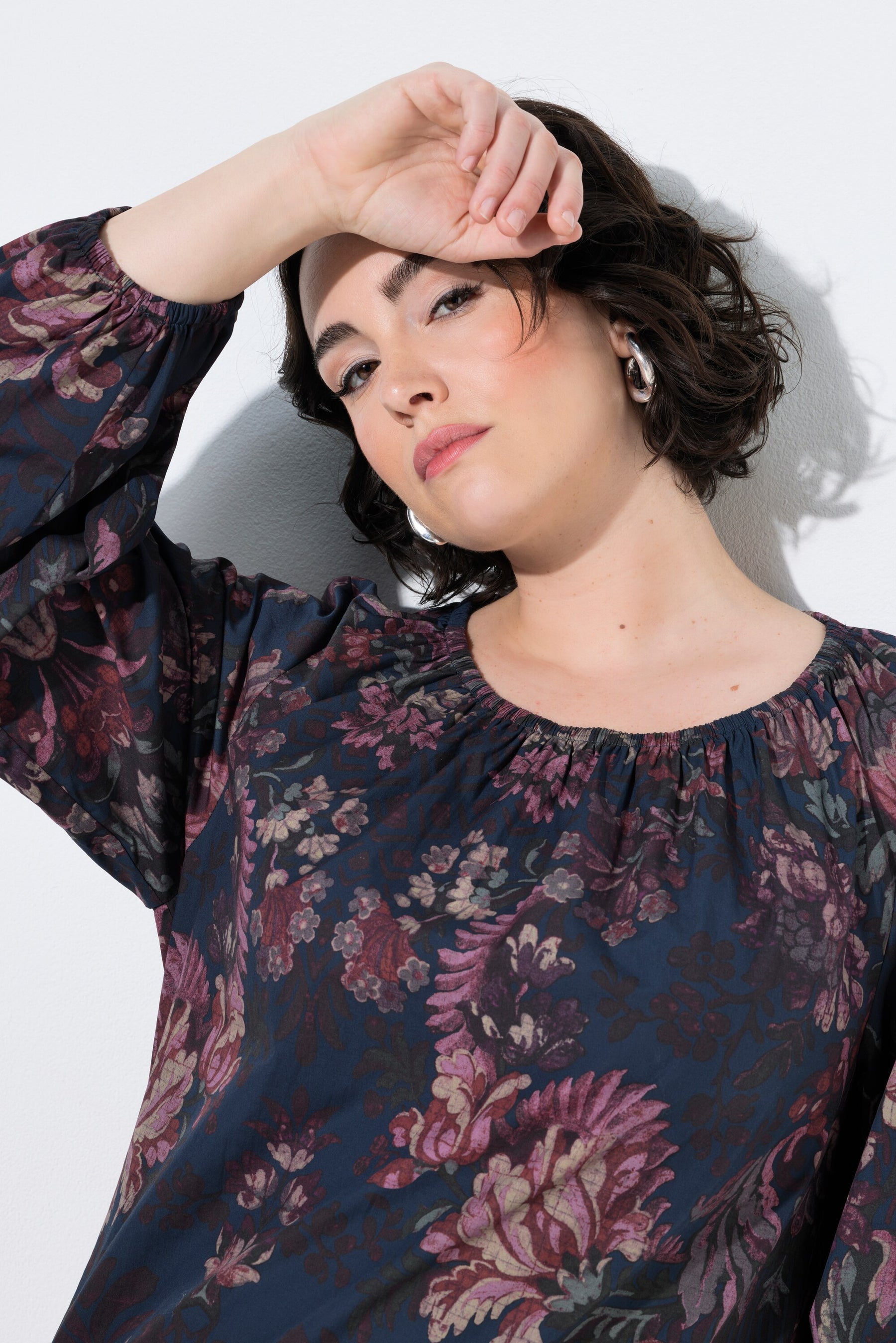A woman wearing the Ulla Popken Floral Damask Blouse in Navy, a dark navy floral blouse with pink and purple flowers, posing against a white wall with her hand near her face.